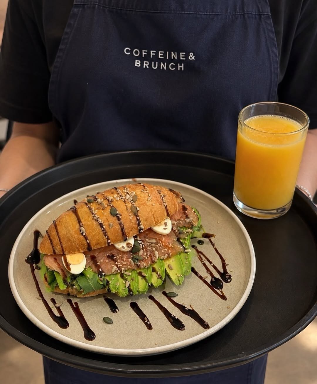 Croissant recheado com salmão, abacate e ovos de codorniz num prato, com um copo de sumo de laranja, num tabuleiro escuro