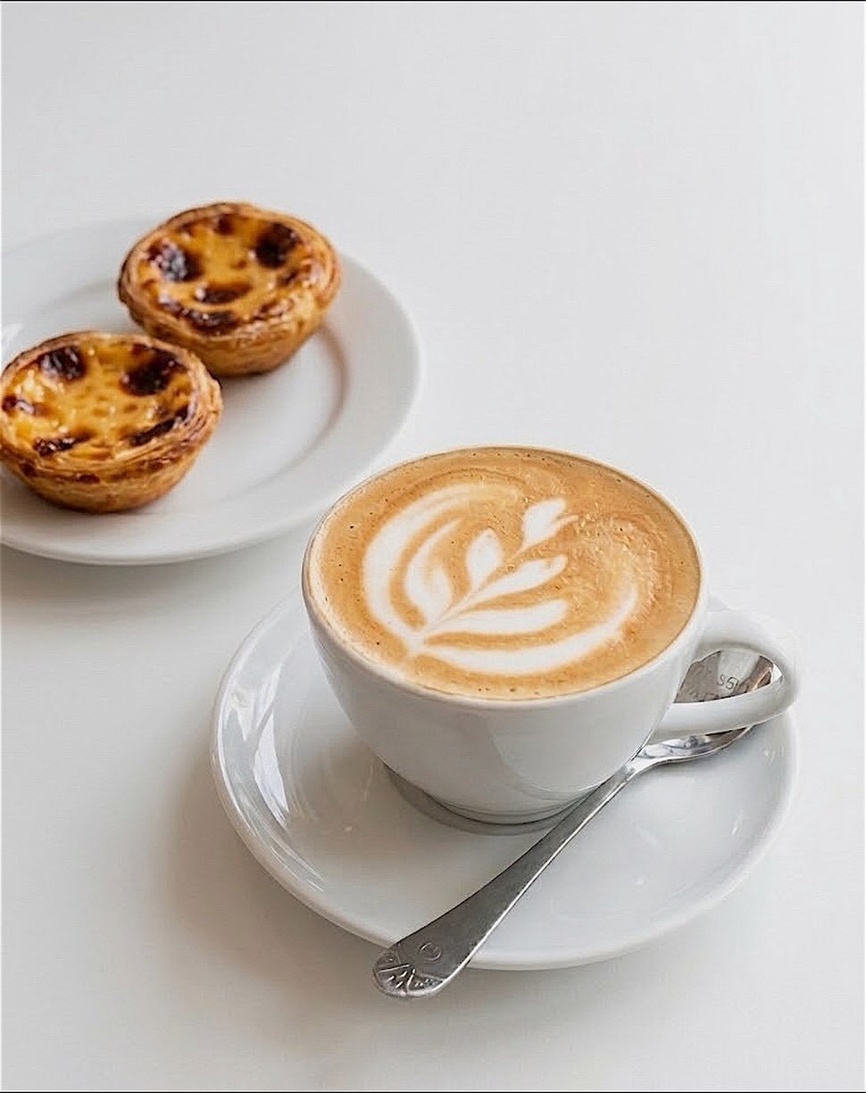 Café com arte no leite numa chávena branca e dois pastéis de nata num prato