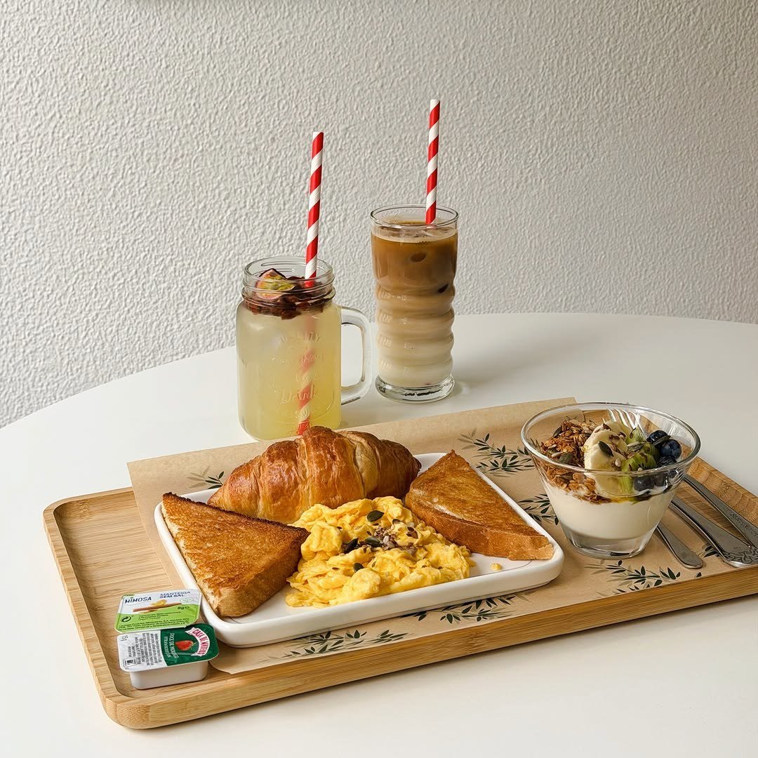 Tabuleiro de madeira com croissant, ovos mexidos, torradas e uma taça de iogurte com granola, com dois copos com palhinhas às riscas